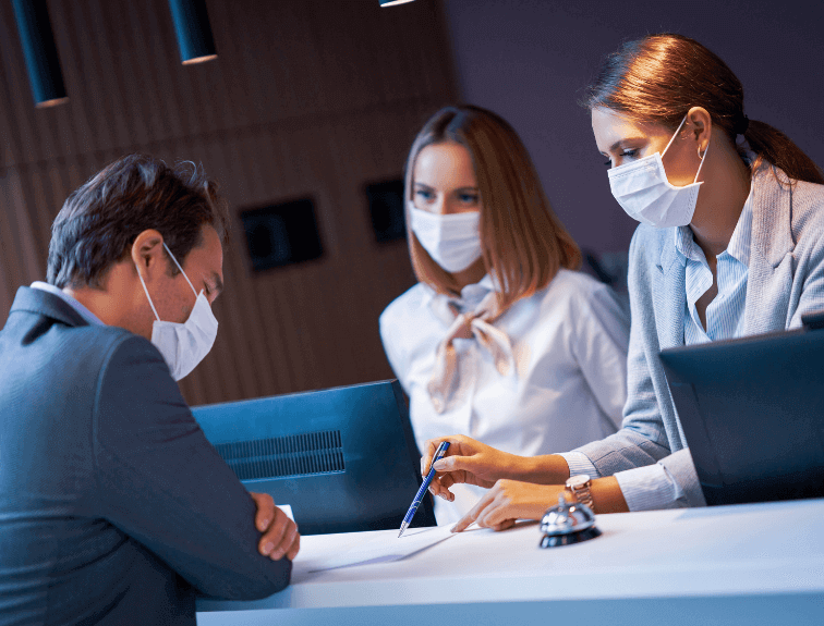 Two concierges wearing masks at hotel desk helping guest wearing mask due to COVID-19 effect on hospitality industry