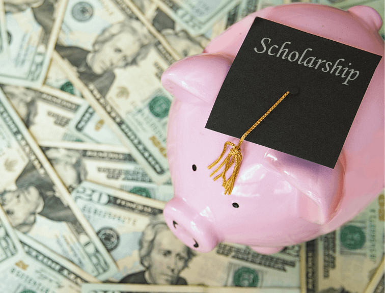 Image of pink piggy bank stand on top of many twenty dollar bills and wearing a college cap with the word scholarships on it